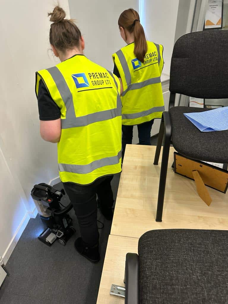 Workers wearing high-viz jackets with the Premac branding work to clean an office space with a vacuum cleaner and cleaning cloths