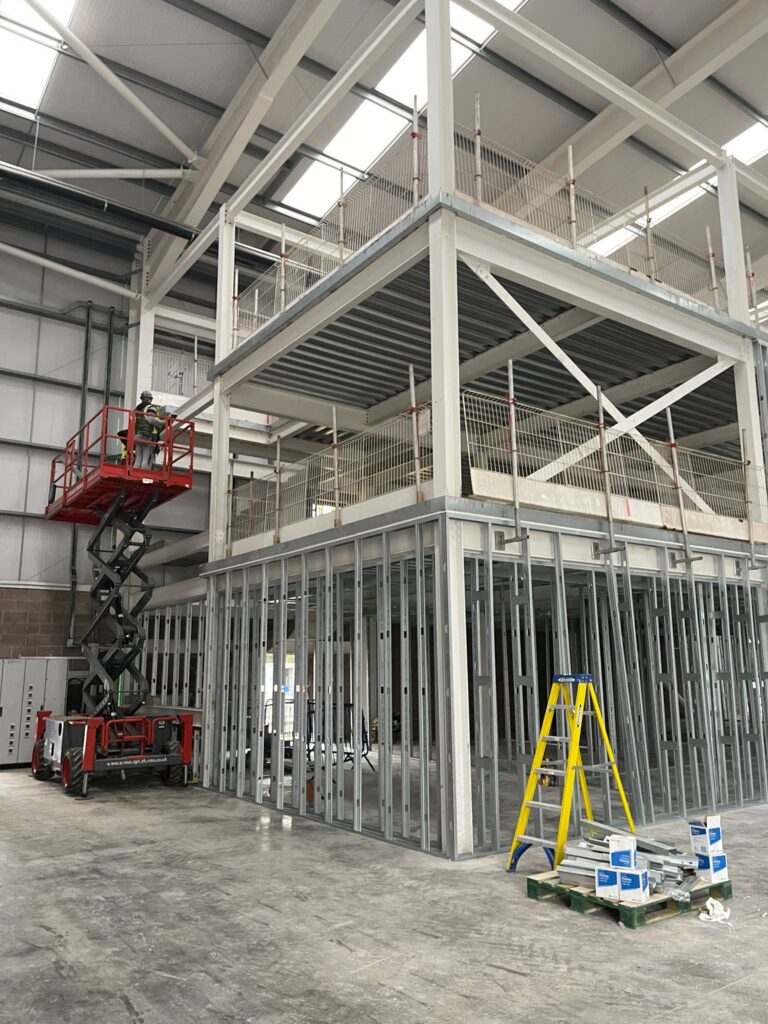 A worker on a cherry picker is building a steel-frame structure inside a warehouse